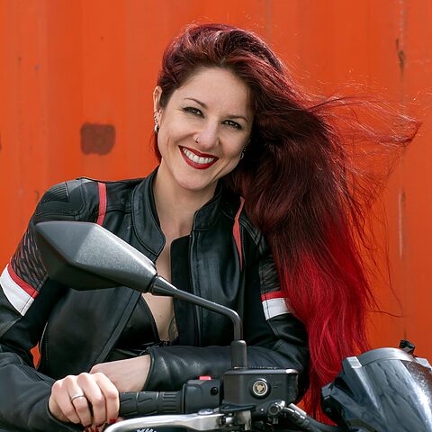 Julia Demann, freie Traurednerin für Traurede NRW, sitzt lachend auf einem Kawasaki-Motorrad vor einer orangefarbenen Wand.