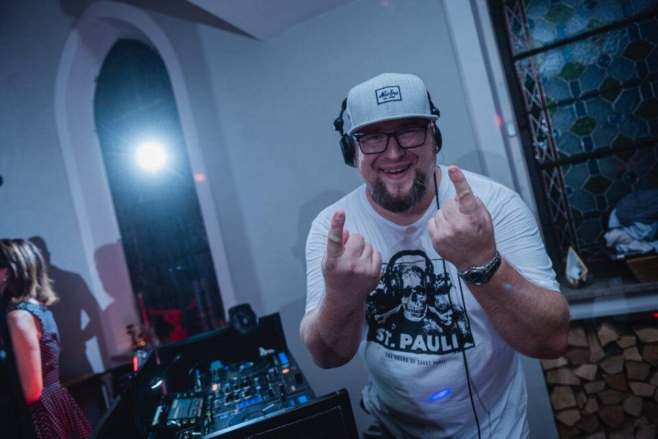 Tom Nawa grinst am DJ-Pult mit Kopfhörern und Rockgeste – alternativer Hochzeits-DJ für Paare, die Rock, Metal und Crossover feiern.