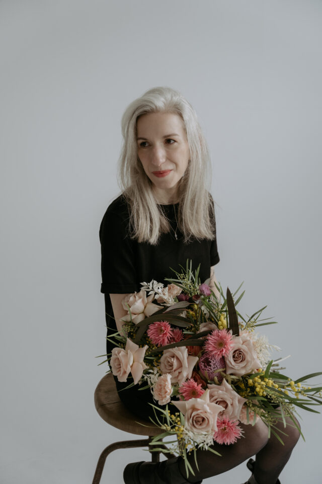 Klara von Eventfloristik by Klara-Elisabeth sitzt vor einer hellgrauen Wand und hält ein großes, locker gebundenes Blumenarrangement auf dem Schoß. Der üppige Strauß besteht aus rosafarbenen und cremefarbenen Rosen, pinken Blüten, gelben Akzenten sowie langen grünen Blättern und Zweigen.