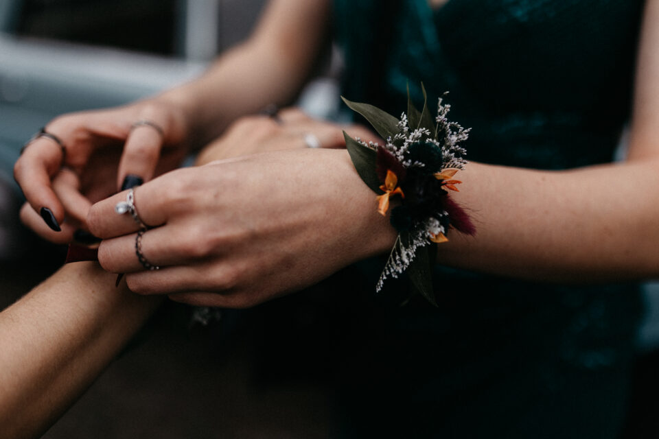Detailaufnahme der Hände einer Person mit schwarzem Nagellack und mehreren filigranen Ringen. Am Handgelenk trägt sie ein kleines florales Armband aus dunklen Blüten, orangefarbenen Akzenten, zarten hellen Blütenrispen und grünen Blättern.