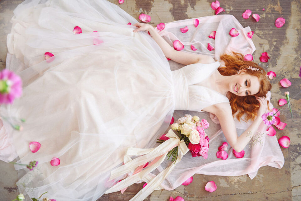 Eine rothaarige Braut liegt lächelnd in einem weit ausgebreiteten weißen Tüllkleid auf dem Boden. Um sie herum und auf dem Kleid liegen zahlreiche pinke Rosenblätter. Neben ihr liegt ein Brautstrauß aus cremefarbenen und kräftig pinken Rosen mit langen hellen Bändern.