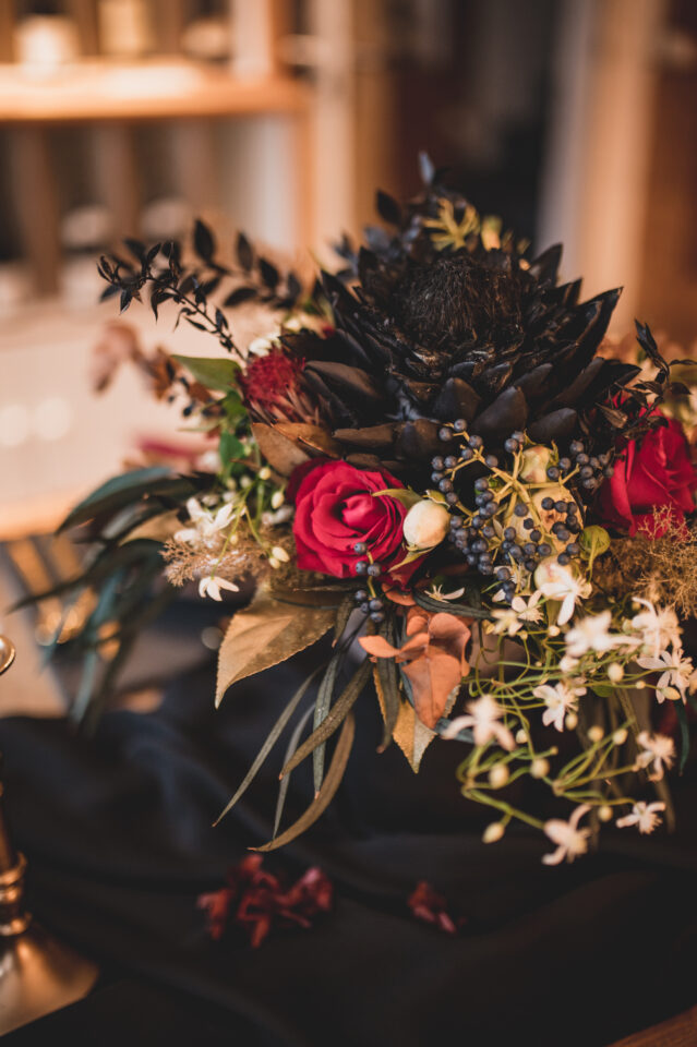 Nahaufnahme eines außergewöhnlich dunklen Blumenarrangements. Im Mittelpunkt steht eine große fast schwarze Blüte, umgeben von tiefroten Rosen, dunklem Blattwerk, blaugrauen Beeren, kleinen weißen Blüten und einzelnen braunen und grünen Akzenten. Die Floristik wirkt üppig, wild und kontrastreich.