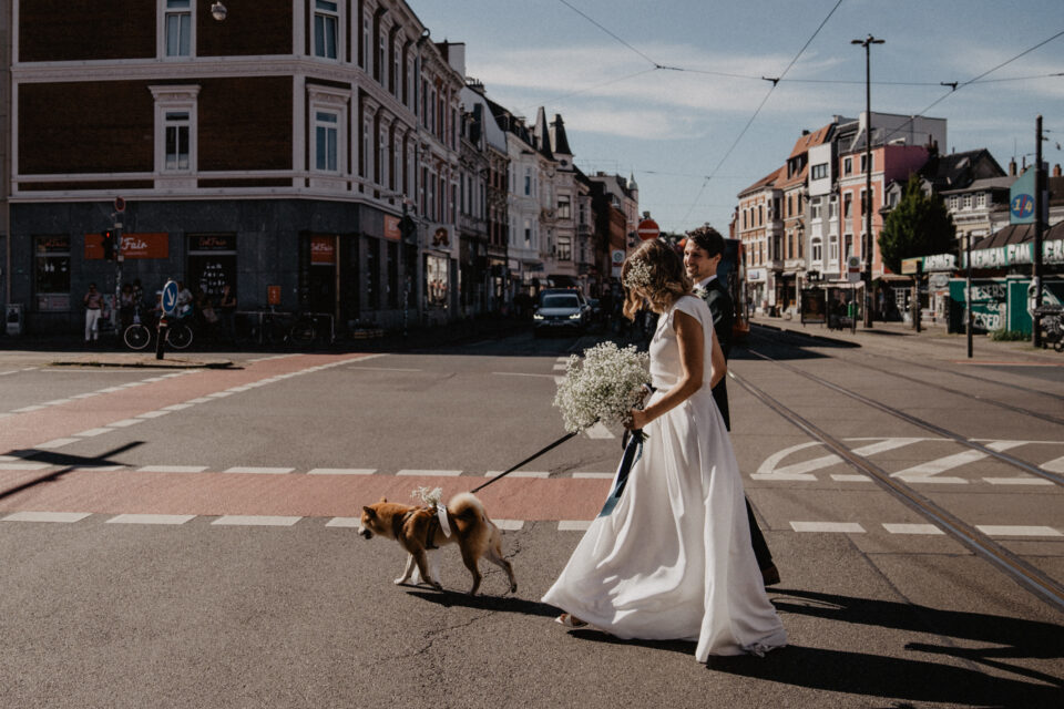 Ein Hochzeitspaar überquert mit einem kleinen braunen Hund an der Leine eine sonnige Straße mit Straßenbahnschienen. Eine Person trägt ein langes weißes Hochzeitskleid und hält einen großen runden Strauß aus weißem Schleierkraut, die andere einen dunklen Anzug. Im Hintergrund stehen mehrstöckige Altbauten und Geschäfte.