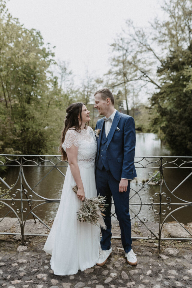 Ein Hochzeitspaar steht auf einer kleinen Steinbrücke vor einem See und schaut sich lächelnd an. Eine Person trägt ein weißes Hochzeitskleid mit Spitzenoberteil und hält einen locker gebundenen Strauß aus getrockneten Gräsern und natürlichen Pflanzenelementen nach unten. Die andere Person trägt einen dreiteiligen blauen Anzug mit Fliege und weißen Sneakern.