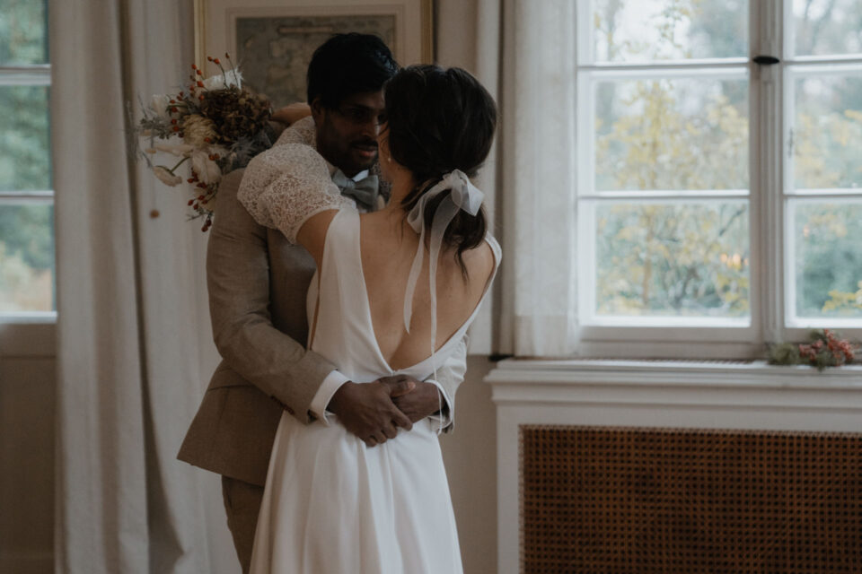 Ein Hochzeitspaar umarmt sich vor großen hellen Fenstern in einem historischen Raum. Eine Person trägt einen beigefarbenen Anzug, die andere ein weißes Hochzeitskleid mit tiefem Rückenausschnitt, kurzen Spitzenärmeln und einer großen hellen Schleife im dunklen Haar. Über der Schulter ist ein locker gebundener Brautstrauß in natürlichen Creme-, Beige- und warmen Rottönen zu sehen.