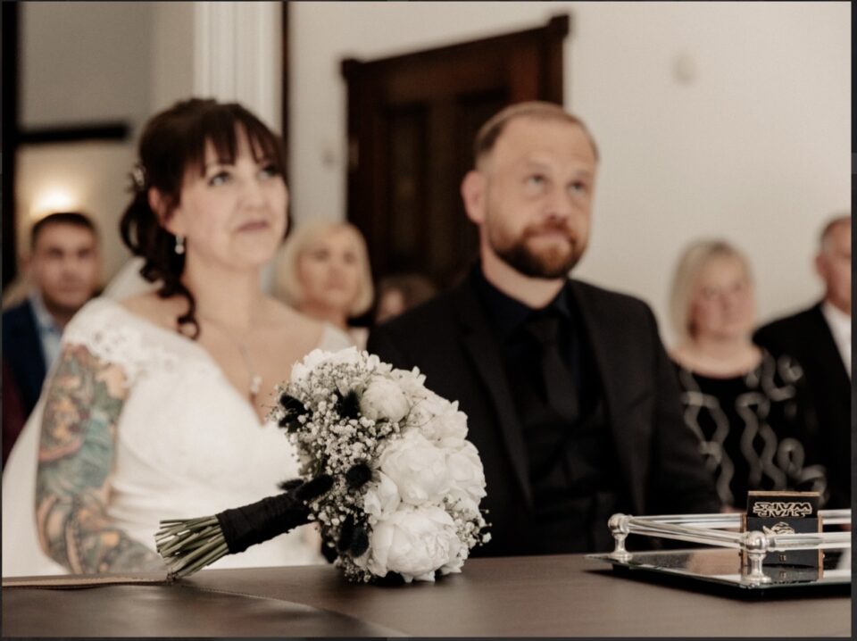 Ein Hochzeitspaar sitzt während der Trauung nebeneinander an einem Tisch. Eine Person trägt ein weißes Hochzeitskleid mit Spitze und zeigt großflächige farbige Tattoos am Arm, die andere einen komplett schwarzen Anzug. Im Vordergrund liegt ein runder Brautstrauß aus großen weißen Blüten, Schleierkraut und kleinen schwarzen floralen Akzenten mit schwarz umwickelten Stielen.