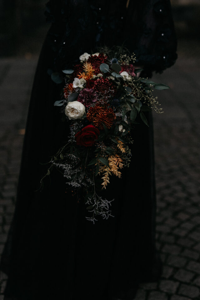 Nahaufnahme eines üppigen, länglich gebundenen Brautstraußes vor einem schwarzen Hochzeitskleid. Dunkelrote Rosen und weitere Blüten in Bordeaux, Rostorange und Creme werden mit Eukalyptus, feinen Zweigen und herabhängendem Blattwerk zu einem wilden, asymmetrischen Arrangement kombiniert.