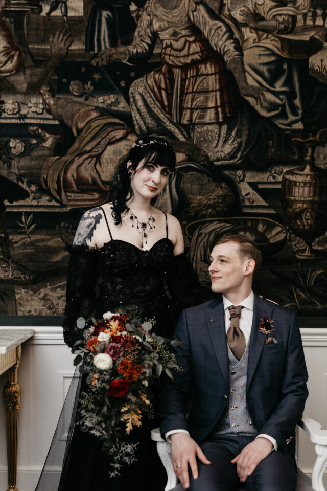 Ein alternativ gekleidetes Hochzeitspaar posiert vor einem großen historischen Wandteppich. Eine Person trägt ein schwarzes, bodenlanges Hochzeitskleid mit Spitze, Tattoos und dunklem Haarschmuck und hält einen üppigen Brautstrauß in Rot, Creme, Orange und dunklem Grün. Daneben sitzt die zweite Person in einem dunkelblauen Anzug mit Weste und passendem floralen Anstecker.