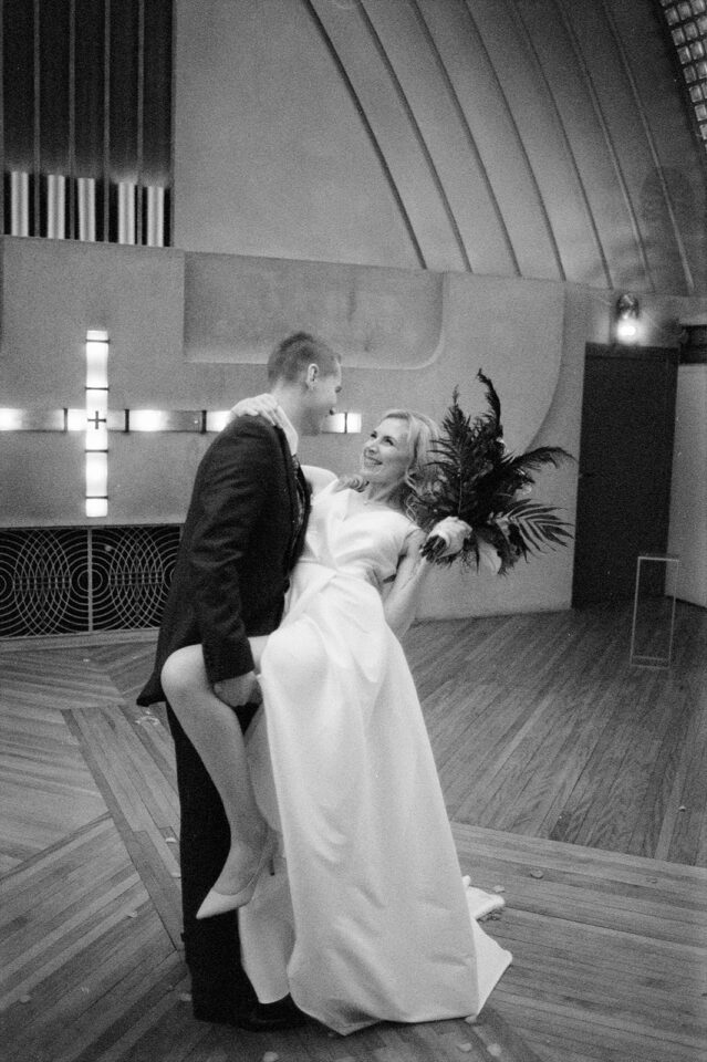 Schwarz-Weiß-Aufnahme eines Hochzeitspaares auf einer großen Tanzfläche mit geometrischem Holzboden. Eine Person im dunklen Anzug hält die andere im langen weißen Hochzeitskleid auf dem Arm. Beide schauen sich lachend an. Die Person im Kleid hält seitlich einen auffälligen dunklen Brautstrauß mit langen, fedrigen Blättern.