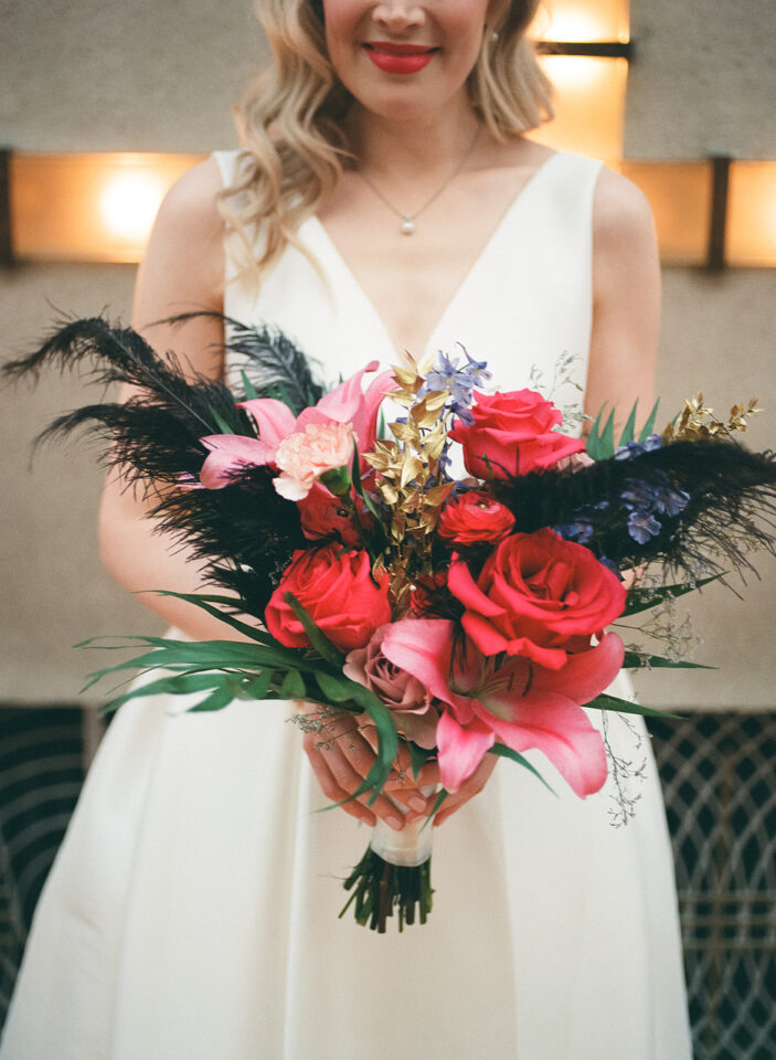 Eine Braut im schlichten weißen Hochzeitskleid hält einen üppigen, farbenfrohen Brautstrauß vor sich. Rote Rosen und Ranunkeln treffen auf große pinke Blüten, violette Akzente, goldfarbenes Blattwerk, lange grüne Blätter und auffällige schwarze Federn. Der Oberkörper der Braut ist zu sehen, ihr Gesicht nur teilweise.