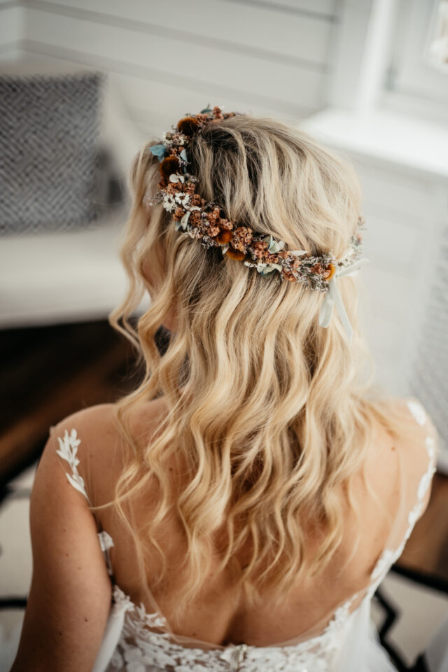 Eine Braut mit langen, welligen blonden Haaren wird von hinten gezeigt. In ihrer halb offenen Hochzeitsfrisur trägt sie einen filigranen Haarkranz aus kleinen getrockneten Blüten, Gräsern und Blättern in Creme, Braun, Rostorange und gedämpftem Grün. Helle Bänder fallen vom Blumenkranz in die Haare; am Rücken ist das transparente, floral bestickte Oberteil ihres Hochzeitskleides zu sehen.