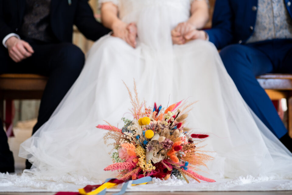 Ein farbenfroher Brautstrauß liegt auf dem ausgebreiteten Rock eines weißen Hochzeitskleides. Das kompakte Arrangement besteht aus Trockenblumen und floralen Elementen in kräftigem Pink, Rot, Orange, Gelb und Blau sowie natürlichen Beige- und Cremetönen. Im unscharfen Hintergrund sitzt das queere Polikül und hält sich an den Händen.