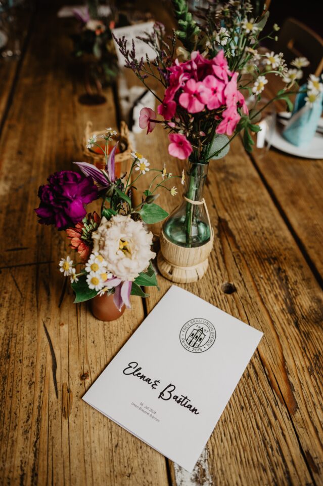 Auf einem rustikalen Holztisch stehen kleine Vasen mit locker arrangierten Blumen in Pink, Violett, Weiß und Orange sowie kleinen weißen Blüten. Davor liegt ein weißes Heft mit den Namen „Elena & Bastian“. Weitere Vasen und Teile der Tischdekoration sind unscharf im Hintergrund zu sehen.