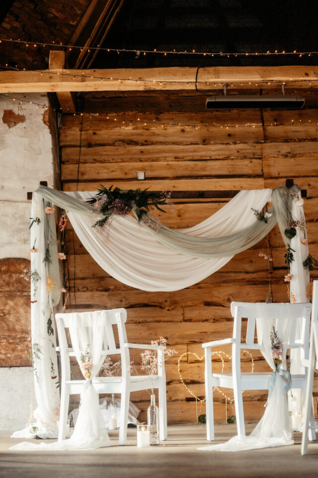 Eine Traukulisse in einem rustikalen Raum mit Holzwänden und sichtbaren Deckenbalken. Zwei weiße Holzstühle stehen vor einem rechteckigen Traubogen, der mit locker drapiertem weißem Stoff, kleinen Blumen, Gräsern und grünen Zweigen dekoriert ist. In der Mitte des Bogens befindet sich ein größeres asymmetrisches Blumenarrangement; Kerzen und Glasvasen stehen am Boden.