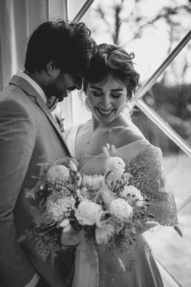 Schwarz-Weiß-Aufnahme eines Hochzeitspaares, das dicht nebeneinander vor einem großen Fenster steht und lachend nach unten schaut. Eine Person trägt einen hellen Anzug, die andere ein Hochzeitskleid mit schulterfreiem Ausschnitt und transparenten Spitzenärmeln. Im Vordergrund hält sie einen großen, locker gebundenen Brautstrauß mit unterschiedlichen Blüten, Gräsern, Beeren und feinem Blattwerk.