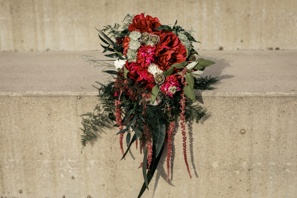 Ein großes asymmetrisches Blumenarrangement liegt vor einer hellen Betonwand. Üppige Blüten in Dunkelrot und kräftigem Pink werden mit weißen Blütendolden, verschiedenen grünen Blättern und langen herabhängenden dunkelroten Blütenständen kombiniert. Das Arrangement läuft nach unten spitz zu und wirft einen deutlichen Schatten auf die Wand.