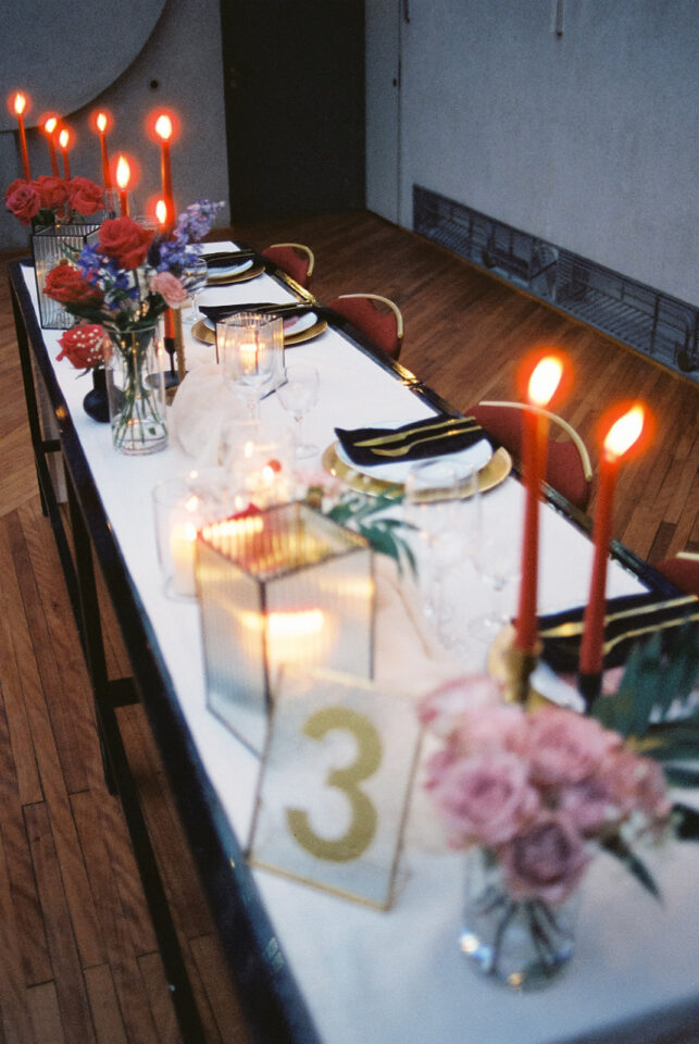 Eine lange festlich gedeckte Hochzeitstafel mit weißer Tischdecke zieht sich durch einen modernen Raum mit dunklem Holzboden. Auf dem Tisch stehen hohe rote Kerzen, leuchtende Windlichter und kleine Blumenarrangements mit roten, pinken und violetten Blüten. Schwarze Servietten, goldene Details und rote Stühle ergänzen die Tischdekoration.