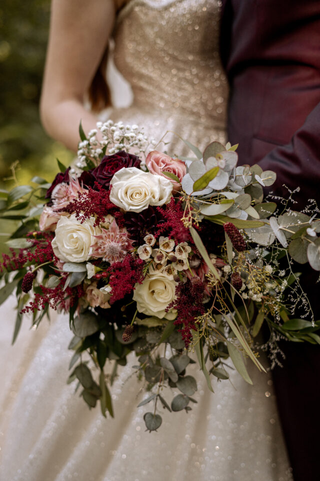 Nahaufnahme eines großen, locker gebundenen Brautstraußes vor einem Hochzeitspaar. Cremeweiße und rosafarbene Rosen werden mit dunkelroten Blüten, kleinen burgunderfarbenen Blütenständen, Eukalyptus und viel locker herabhängendem Grün kombiniert. Im Hintergrund sind ein glitzerndes helles Kleid und ein dunkelroter Anzug zu erkennen.
