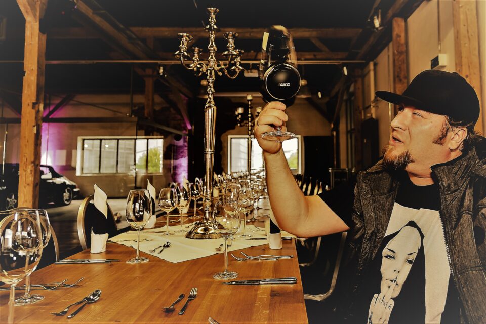 Tom Nawa sitzt in einer Hochzeitslocation an einer festlich gedeckten Tafel und hält Kopfhörer hoch – Hochzeits-DJ für alternative Feiern mit Rock, Metal und eigenem Sound.