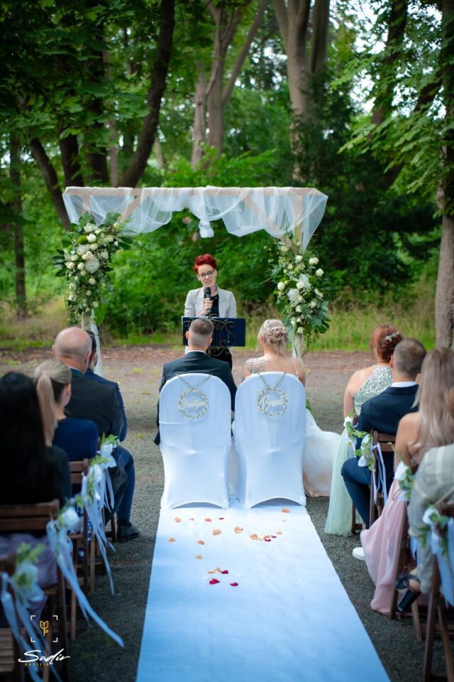 Eine freie Trauung findet draußen unter hohen grünen Bäumen statt. Das Hochzeitspaar sitzt mit dem Rücken zur Kamera auf zwei weißen Stühlen vor einem mit weißem Stoff und weißen Blumen dekorierten Traubogen. Dahinter steht die freie Rednerin Mareike mit Mikrofon am Rednerpult. Auf beiden Seiten des weißen Mittelgangs sitzen die Hochzeitsgäste.