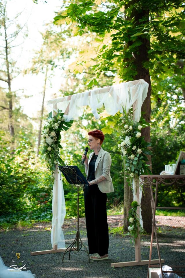 Freie Rednerin Mareike steht bei einer Trauung im Grünen unter einem mit weißem Stoff und großen weißen Blumenarrangements dekorierten Traubogen. Sie trägt einen hellen Blazer über schwarzer Kleidung, hat kurze rote Haare und spricht mit einem Mikrofon hinter einem geschwungenen Rednerpult.