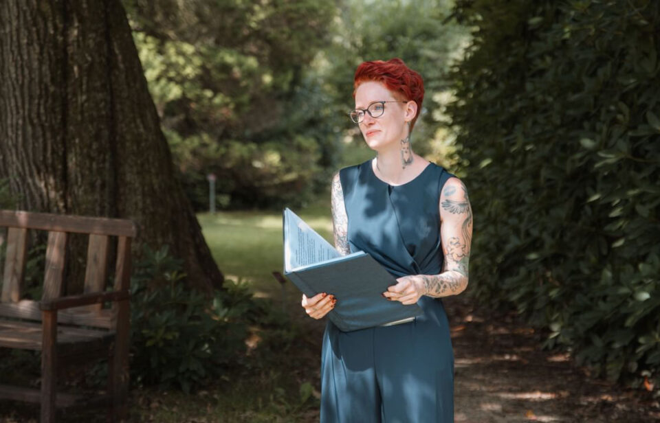 Freie Rednerin Mareike steht mit einer geöffneten Mappe in den Händen auf einem schattigen Weg im Grünen. Sie trägt eine dunkelblaue ärmellose Kombination und eine Brille, hat kurze rote Haare sowie zahlreiche Tattoos an Armen und Hals.