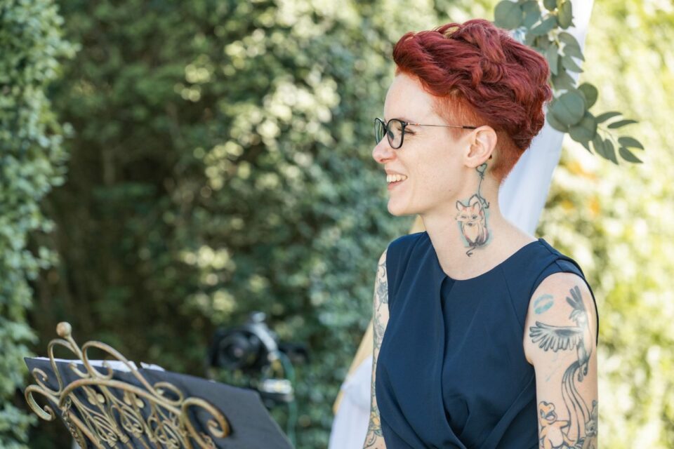 Freie Rednerin Mareike steht während einer Trauung im Freien und lächelt zur Seite. Sie trägt eine dunkelblaue ärmellose Kombination und eine Brille, hat kurze rote Haare und zahlreiche farbige Tattoos an Armen und Hals. Vor ihr steht ein geschwungenes metallenes Rednerpult.