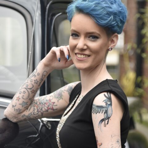Portrait von Mareike mit kurzen leuchtend blauen Haaren, die lächelnd neben einem dunklen Fahrzeug steht. Sie trägt ein schwarzes ärmelloses Oberteil und ist an beiden Armen großflächig bunt tätowiert. Kleine Piercings und Ohrschmuck sind ebenfalls zu sehen.