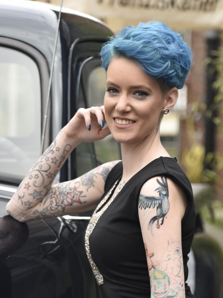 Portrait von Mareike mit kurzen leuchtend blauen Haaren, die lächelnd neben einem dunklen Fahrzeug steht. Sie trägt ein schwarzes ärmelloses Oberteil und ist an beiden Armen großflächig bunt tätowiert. Kleine Piercings und Ohrschmuck sind ebenfalls zu sehen.