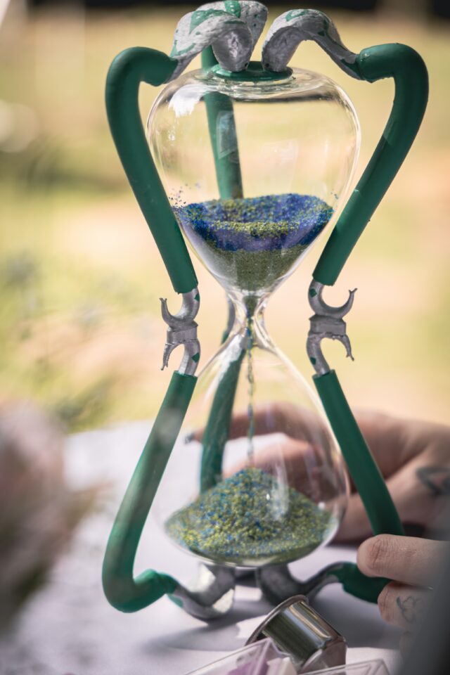 Nahaufnahme einer gläsernen Sanduhr mit einem auffälligen grünen Gestell. Blau-grüner Sand rieselt durch die schmale Mitte in die untere Hälfte, während eine Hand die Sanduhr festhält.
