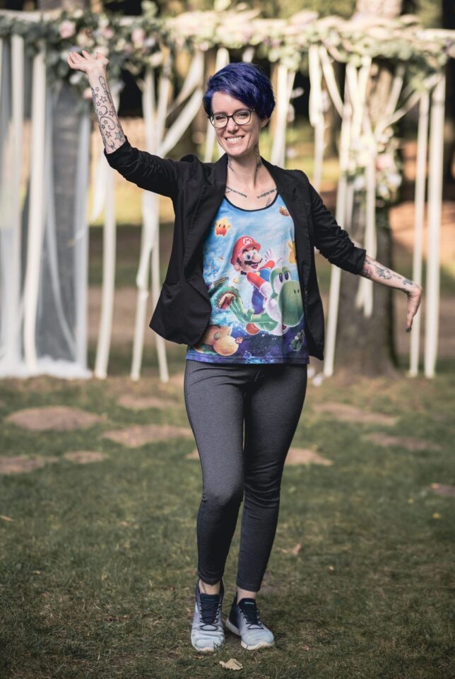 Mareike steht lächelnd auf einer Wiese und hebt einen Arm in die Luft. Sie hat kurze blaue Haare, trägt eine schwarze Jacke, dunkle Hose und ein buntes Shirt mit Super Mario und Yoshi. An beiden Armen sind zahlreiche Tattoos zu sehen.