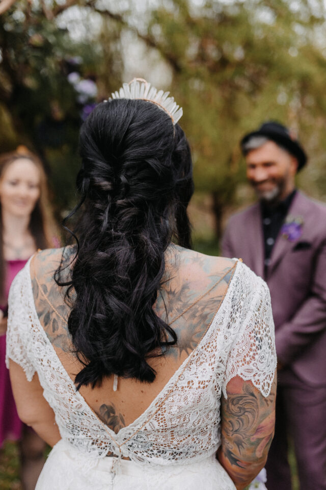 Heiratende mit kunstvoller Rückenansicht in Spitzenkleid und Kristallkrone bei einer freien Trauung im Grünen – Stil trifft Tattoo.