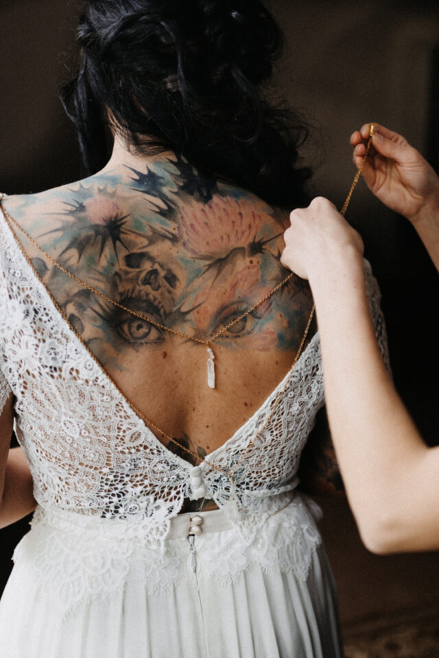 Detailaufnahme beim Getting Ready: Kristallkette wird über tätowierten Rücken der Braut mit Spitzenkleid gelegt.