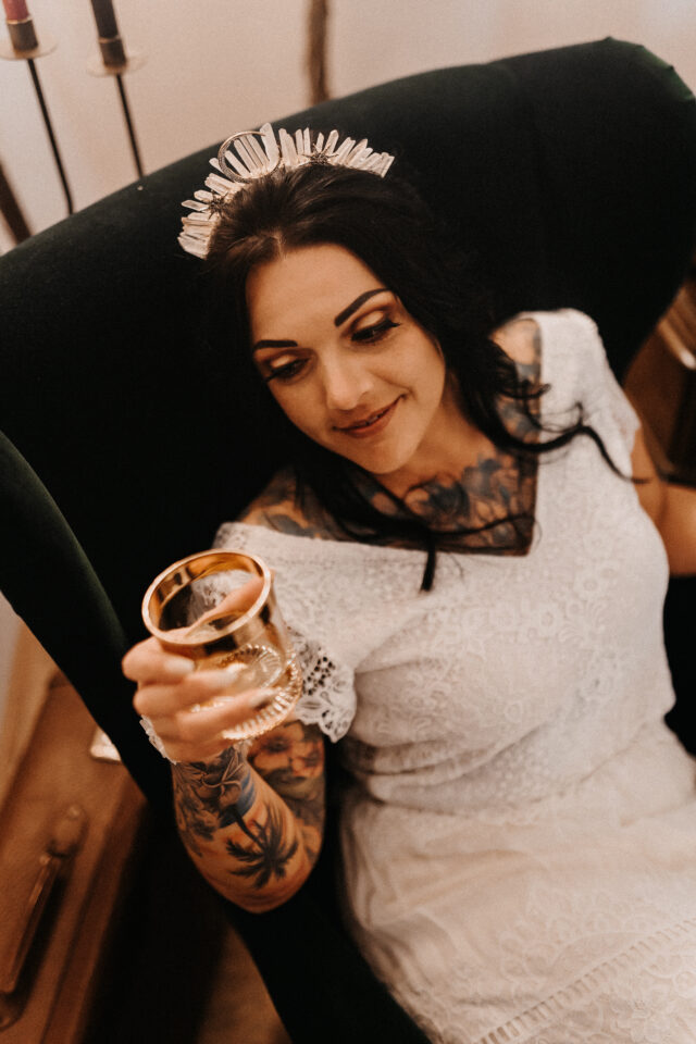 Braut in Spitzenkleid mit Kristallkrone lehnt entspannt im Sessel und hebt ein Glas – alternative Hochzeitsvibes im mystischen Metal-Style pur.