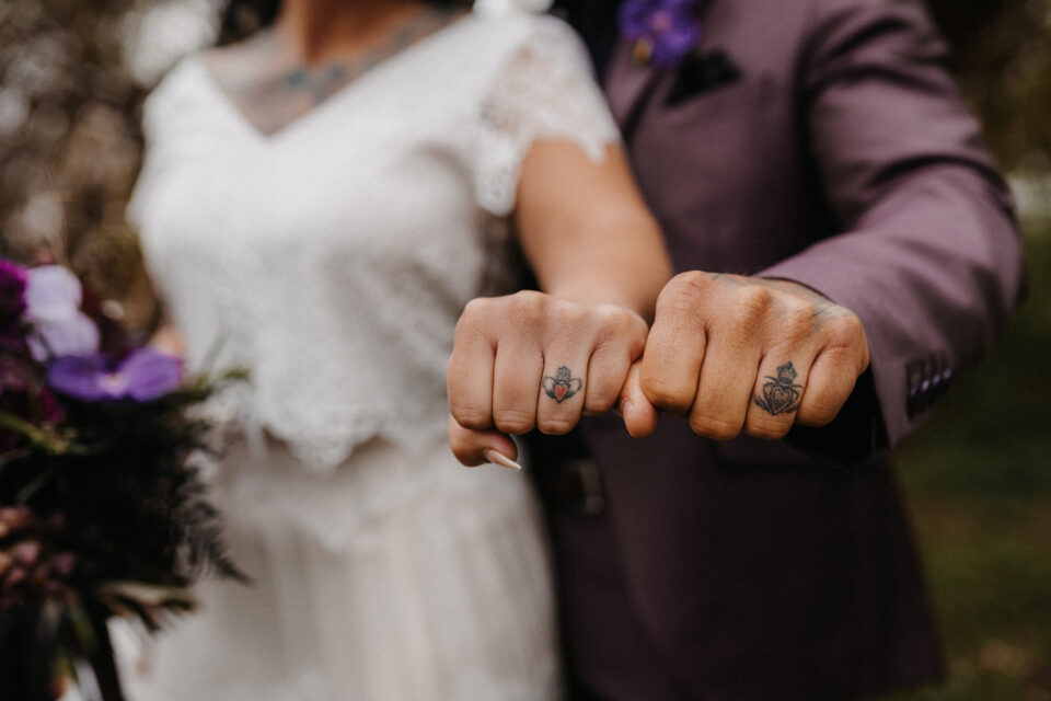 Hochzeitspaar mit Tattoos an den Ringfingern zeigt geballte Fäuste – alternative Hochzeitsinspiration mit individuellen Liebessymbolen