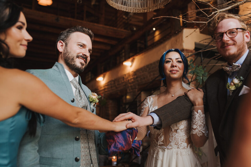 Während einer Trauung reichen sich zwei Personen die Hände. Eine Person im graugrünen Anzug hält die Hand einer Person in einem türkisfarbenen Kleid, während eine Braut im weißen Spitzenkleid und ein Mann im dunklen Anzug danebenstehen und die Szene beobachten. Die Zeremonie findet in einem rustikalen Innenraum mit Holzbalken und Zweigen als Dekoration statt.