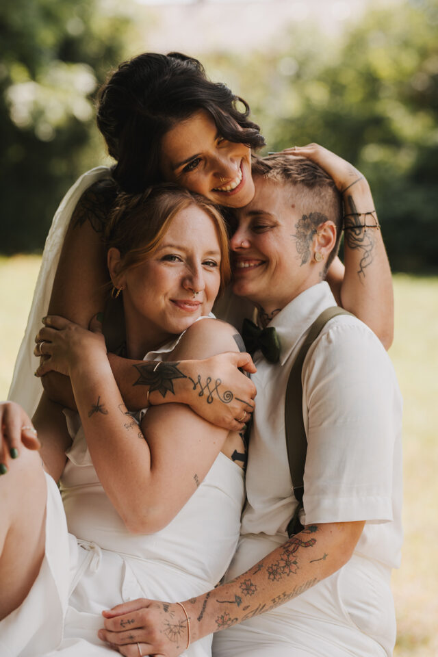 Drei Personen in heller Hochzeitskleidung sitzen draußen eng aneinander und umarmen sich. Alle drei lächeln in die Kamera oder haben die Augen geschlossen. Zwei Personen sind sichtbar tätowiert; eine trägt ein weißes Hemd mit schwarzer Fliege und Hosenträgern. Im Hintergrund ist eine sonnige Grünfläche zu sehen.