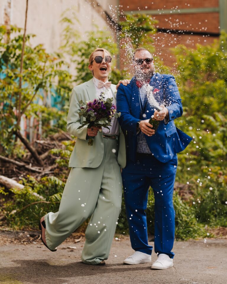 Ein Hochzeitspaar steht lachend im Freien zwischen grünen Pflanzen und trägt Sonnenbrillen. Eine Person im hellgrünen Hosenanzug mit rosa Fliege hält einen Brautstrauß aus violetten und grünen Blumen. Die andere Person im leuchtend blauen Anzug öffnet eine Flasche, aus der Schaum und Tropfen durch die Luft spritzen.