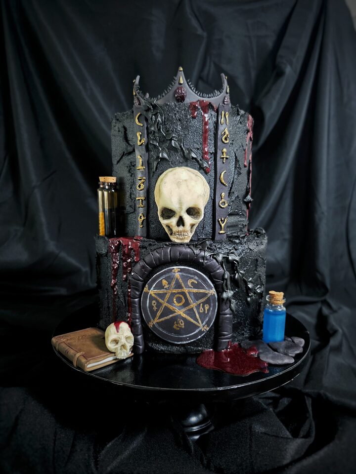 Zweistöckige schwarze Motivtorte im düsteren Gothic-Stil mit Totenschädel, Krone, goldenen Symbolen, dunkelroten tropfenden Details und einem Pentagramm auf der Vorderseite. Neben der Torte stehen kleine Fläschchen, ein Buch und ein weiterer Totenschädel.