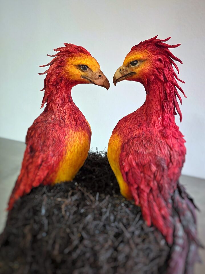 Zwei detailreich modellierte rote und orangefarbene Phönixe sitzen einander zugewandt auf einem dunklen Nest. Ihre Köpfe neigen sich zueinander, sodass zwischen ihren Schnäbeln die Form eines Herzens entsteht.