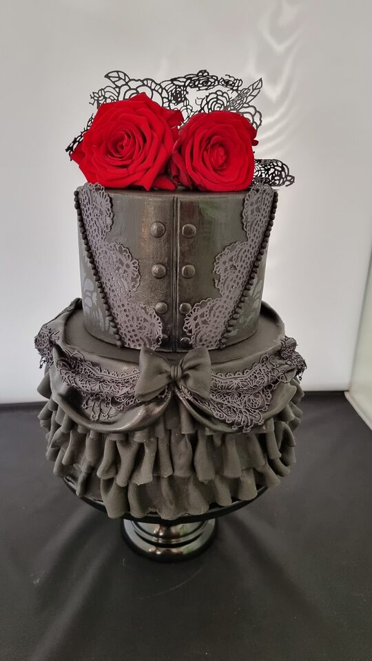 Zweistöckige dunkelgraue Hochzeitstorte im Gothic-Stil. Die untere Etage ist mit üppigen Rüschen, Spitzenornamenten und einer Schleife dekoriert, die obere erinnert mit Knöpfen und Spitzenelementen an ein Korsett. Zwei leuchtend rote Rosen und filigrane schwarze Blütendekoration bilden einen starken Kontrast.