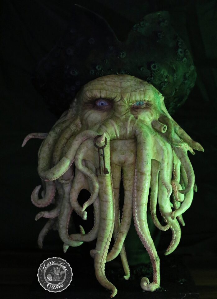 Detailreiche Tortenskulptur von Davy Jones mit zahlreichen langen Tentakeln im Gesicht, bläulich-grünen Augen und seinem charakteristischen dunklen Hut. Zwischen den Tentakeln hängt ein alter Schlüssel. Die Figur ist grünlich beleuchtet und steht vor schwarzem Hintergrund.