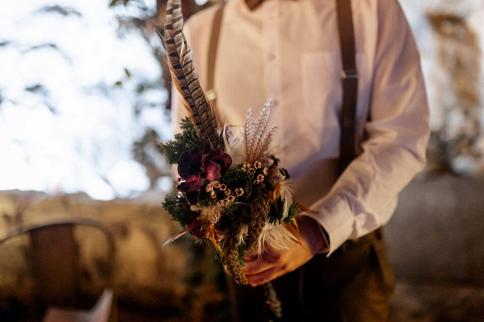 Eine Person in weißem Hemd, brauner Fliege und Hosenträgern hält einen kleinen, ungewöhnlichen Blumenstrauß vor sich. Dunkelrote und violette Blüten, Grün, getrocknete Gräser und Pflanzenelemente werden mit mehreren langen braun gemusterten Federn und kleineren hellen Federn kombiniert.