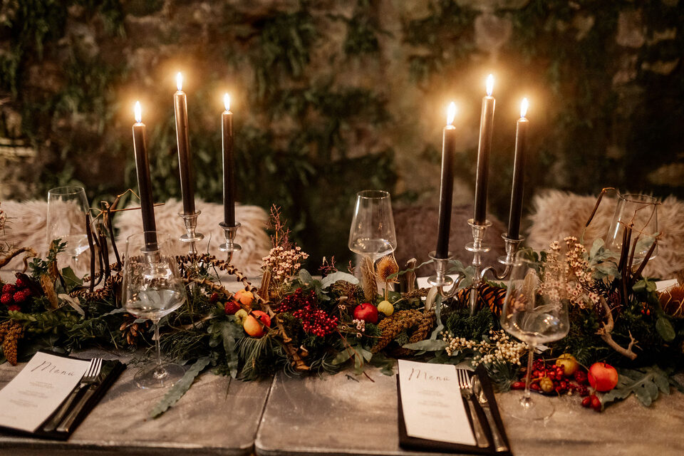 Eine dunkel und herbstlich dekorierte Hochzeitstafel ist mit schwarzen Stabkerzen in gläsernen Kerzenhaltern, Weingläsern und dunklem Geschirr eingedeckt. Über die Tischmitte zieht sich ein üppiges Arrangement aus tiefgrünem Blattwerk, roten Beeren, kleinen Früchten, Zweigen sowie Blüten und Pflanzenelementen in Rot, Braun und Creme. Helle Felle liegen auf den Sitzplätzen.