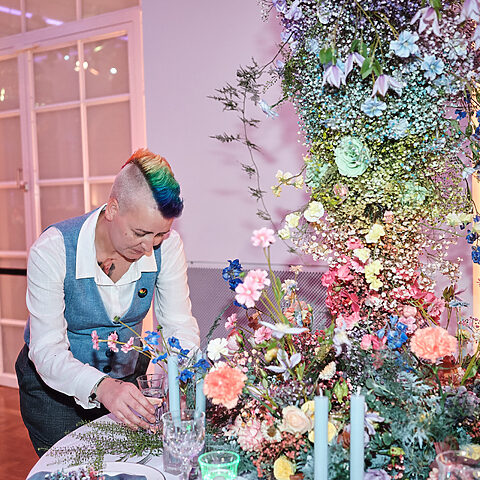 Floristin Schmi von Die Blumenfee steht an einer aufwendig dekorierten Hochzeitstafel und richtet Gläser und florale Details her. Sie trägt Hemd und Weste und hat einen kurzen, seitlich rasierten Irokesenschnitt in Regenbogenfarben. Vor und neben ihr wachsen üppige Blumenarrangements in Rosa, Blau, Gelb und Weiß über den Tisch und weit in die Höhe.