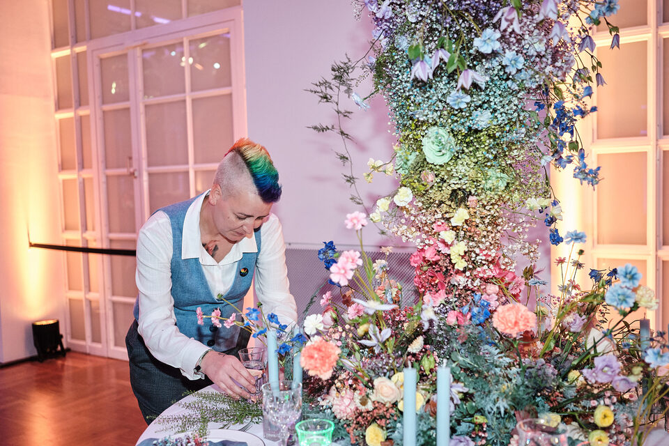 Floristin Schmi von Die Blumenfee steht an einer aufwendig dekorierten Hochzeitstafel und richtet Gläser und florale Details her. Sie trägt Hemd und Weste und hat einen kurzen, seitlich rasierten Irokesenschnitt in Regenbogenfarben. Vor und neben ihr wachsen üppige Blumenarrangements in Rosa, Blau, Gelb und Weiß über den Tisch und weit in die Höhe.