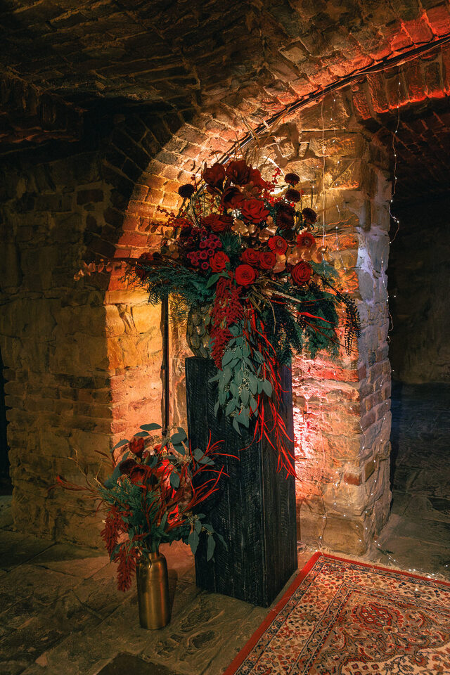 Eine große florale Installation steht in einem dunklen historischen Steingewölbe vor einer von warmem Licht angestrahlten Backsteinwand. Das Arrangement besteht aus zahlreichen roten und dunkelroten Blüten, tiefgrünem Blattwerk, Eukalyptus sowie langen herabhängenden roten Zweigen und Pflanzenelementen. Ein kleineres Arrangement in ähnlichen Farben steht daneben in einer hohen goldfarbenen Vase.