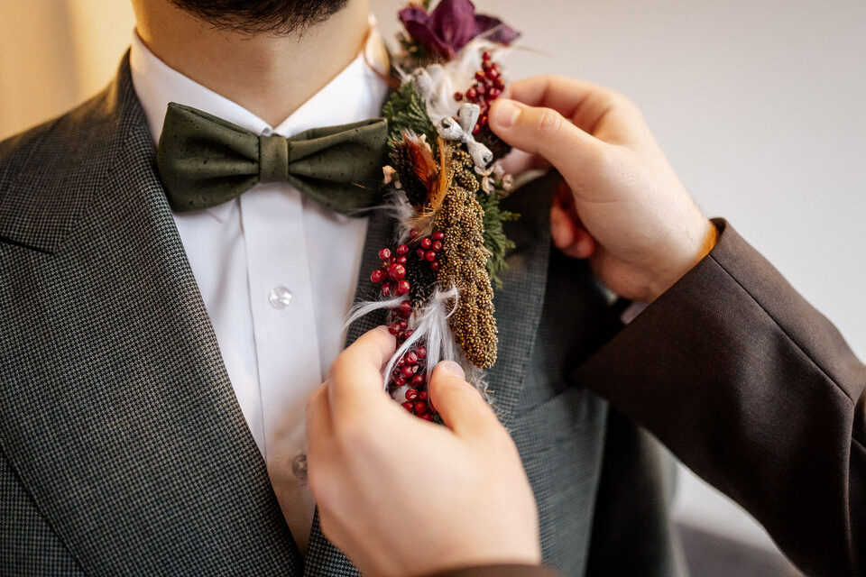 Nahaufnahme eines Bräutigams im dunkelgrauen Anzug mit weißem Hemd und dunkelgrüner Fliege. Eine Person befestigt an seinem Revers einen auffälligen floralen Anstecker aus dunkelroten Blüten, roten Beeren, kleinen Zweigen, Federn und verschiedenen getrockneten Pflanzenelementen.