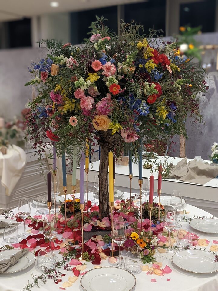 Eine runde Hochzeitstafel ist mit einer großen baumartigen Blumeninstallation dekoriert. Über dem Tisch bildet eine üppige Krone aus pinken, roten, gelben, blauen, violetten und cremefarbenen Blüten sowie lockerem Grün ein farbenfrohes Dach. Auf der weißen Tischdecke liegen zahlreiche bunte Blütenblätter; dazwischen stehen hohe schmale Kerzen in verschiedenen Farben und goldfarbenen Haltern.