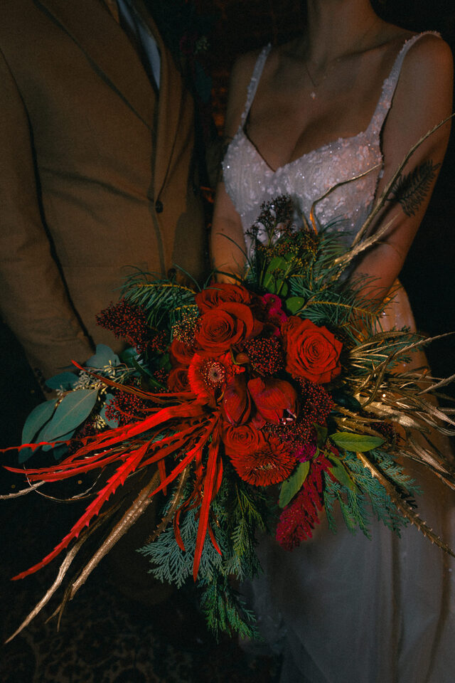 Nahaufnahme eines Hochzeitspaares mit einem großen asymmetrischen Brautstrauß. Kräftig rote und dunkelrote Rosen sowie weitere Blüten werden mit tiefgrünem Blattwerk, Zweigen und langen roten und goldfarbenen Pflanzenelementen kombiniert, die weit aus dem Strauß herausragen. Die Braut trägt ein glitzerndes weißes Kleid, daneben ist eine Person in einem braunen Anzug zu sehen.